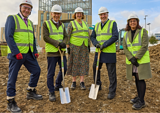 A group of people in hard hats and high vis jackets stand with spades on a construction site 