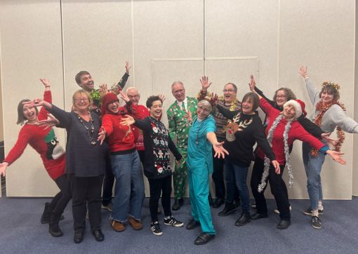 Image of a choir called The Part Singers. They are dressed in tinsel and Christmas jumpers with their hands outstretched